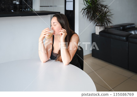 Cute, slightly sleepy young woman enjoying a warm cappuccino at home in a bright cozy kitchen. Simple morning routine, relaxed atmosphere, casual home lifestyle, calm start of the day. 135086769