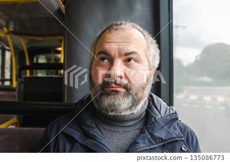 Portrait of an elderly European man sitting inside a tram, calmly waiting for his stop. Casual everyday scene, thoughtful expression, public transport interior, urban atmosphere, soft daylight through Portrait of an elderly European man sitting inside a tram, calmly waiting for his stop. Casual everyday scene, thoughtful expression, public transport interior, urban atmosphere, soft daylight through 135086773