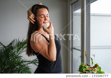 Cute, slightly sleepy young woman enjoying a warm cappuccino at home in a bright cozy kitchen. Simple morning routine, relaxed atmosphere, casual home lifestyle, calm start of the day. Cute, slightly sleepy young woman enjoying a warm cappuccino at home in a bright cozy kitchen. Simple morning routine, relaxed atmosphere, casual home lifestyle, calm start of the day. 135086777