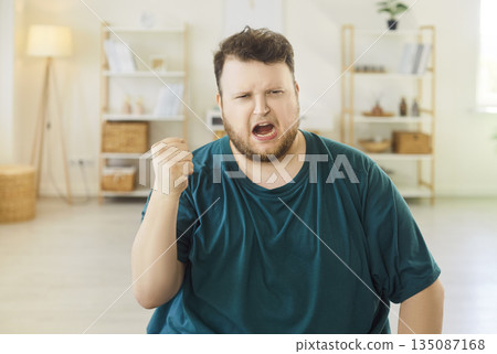 Frustrated man screaming with raised hands at home, expressing stress and rage Frustrated man screaming with raised hands at home, expressing stress and rage 135087168