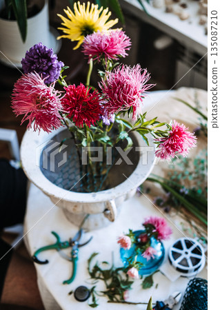 Purple and pink aster flowers stand in wire mesh inside white stone urn on cluttered work table. Creative wellness, stress relief through plants, mindful floral arranging. 135087210