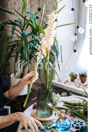 Hands hold tall white gladiolus stems over green glass vase on work table. Zero-waste floristry, sustainable floral foam alternatives, eco-friendly flower arranging. 135087215