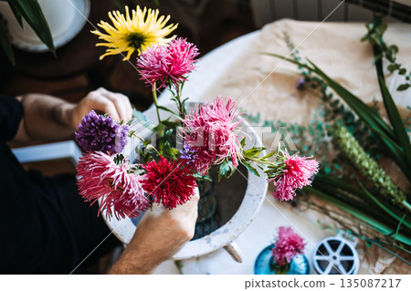 Purple and pink aster flowers stand in wire mesh inside white stone urn on cluttered work table. Creative wellness, stress relief through plants, mindful floral arranging. Purple and pink aster flowers stand in wire mesh inside white stone urn on cluttered work table. Creative wellness, stress relief through plants, mindful floral arranging. 135087217