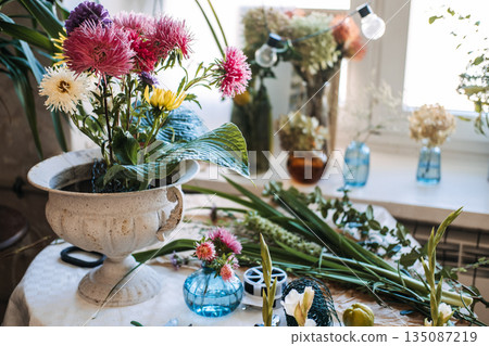 Large green hosta leaves and pink asters sit inside white stone urn on table with floral supplies. Floral design class, DIY flower arranging, floristry workshop, creative education. 135087219