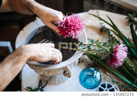 Hands place pink aster flower into wire mesh inside stone urn on work table. Traditional floristry techniques, handcrafted bouquets, authentic creative process. 135087233