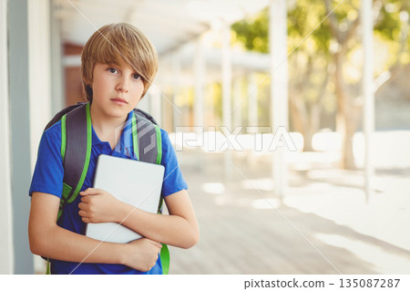 School-age boy holding tablet while standing on school walkway wearing blue polo shirt and backpack School-age boy holding tablet while standing on school walkway wearing blue polo shirt and backpack 135087287