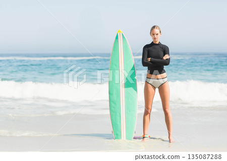 Woman standing on shoreline wearing black rash guard and bikini bottoms beside striped surfboard 135087288