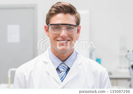Male laboratory researcher standing at lab bench wearing white lab coat and safety goggles 135087298