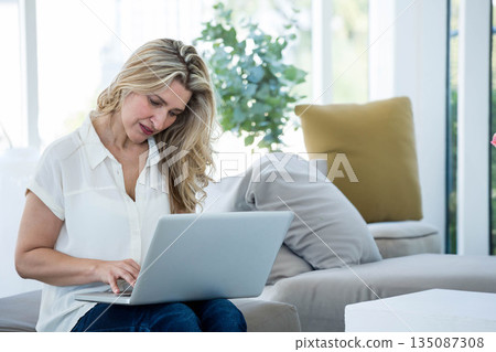 Woman typing on silver laptop while sitting on light-colored sofa under floor-to-ceiling windows 135087308