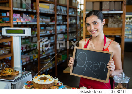 Woman smiling behind bakery counter holding chalkboard Open sign beside cake, copy space 135087311