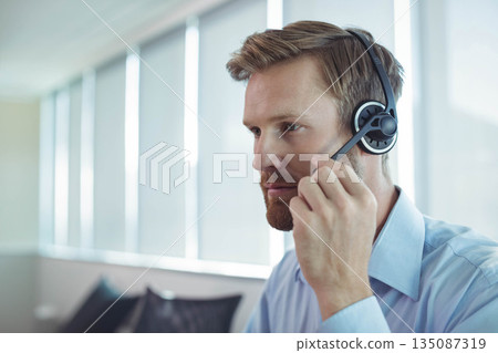 Male office worker wearing dress shirt adjusting headset mic at office desk with blinds, copy space Male office worker wearing dress shirt adjusting headset mic at office desk with blinds, copy space 135087319