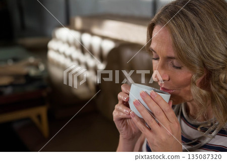 Mature adult woman sitting on sofa sipping white mug beside coffee table in lounge, copy space Mature adult woman sitting on sofa sipping white mug beside coffee table in lounge, copy space 135087320