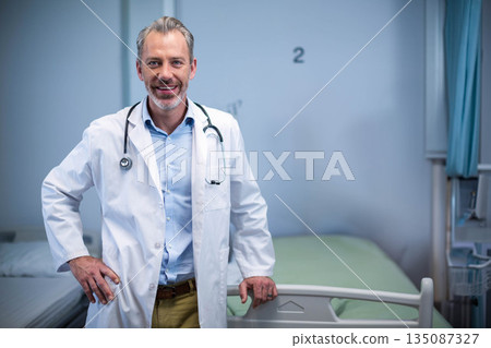 Male doctor wearing lab coat leaning on bed rail in hospital ward with stethoscope, privacy curtain 135087327