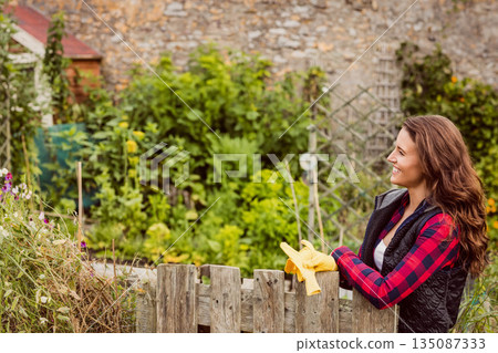 Woman leaning on picket fence in garden with plants holding yellow gardening gloves, copy space Woman leaning on picket fence in garden with plants holding yellow gardening gloves, copy space 135087333