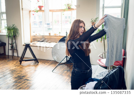 Woman in mid 20s examining gray t-shirt in studio with reflective disc, red chair, copy space Woman in mid 20s examining gray t-shirt in studio with reflective disc, red chair, copy space 135087335