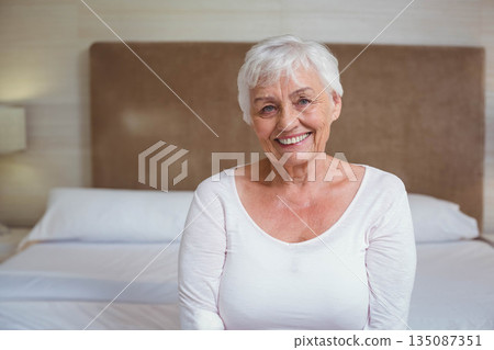 Senior woman sitting on bed edge in bedroom, featuring padded headboard and lamp on bedside table Senior woman sitting on bed edge in bedroom, featuring padded headboard and lamp on bedside table 135087351
