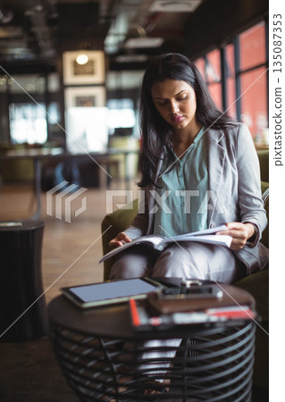 Businesswoman reviewing documents on lounge chair beside table with tablet and notebook at office Businesswoman reviewing documents on lounge chair beside table with tablet and notebook at office 135087353