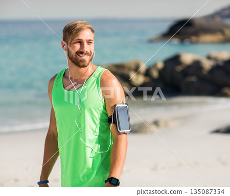 Man wearing tank top, watch, smartphone armband, wired earphones pausing run on beach, copy space 135087354