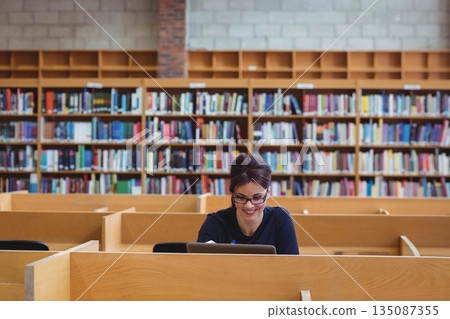 Woman wearing glasses working at study carrel in academic library with laptop, notebook and pen Woman wearing glasses working at study carrel in academic library with laptop, notebook and pen 135087355
