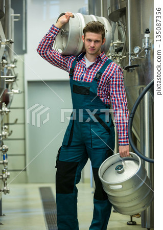 Male brewery technician carrying two kegs in brewery corridor lined with stainless tanks and hoses 135087356