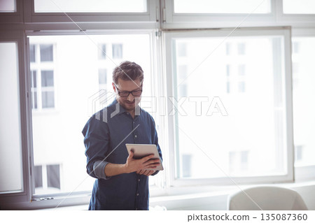 Man wearing glasses and blue button-up shirt standing by window using tablet in office, copy space 135087360