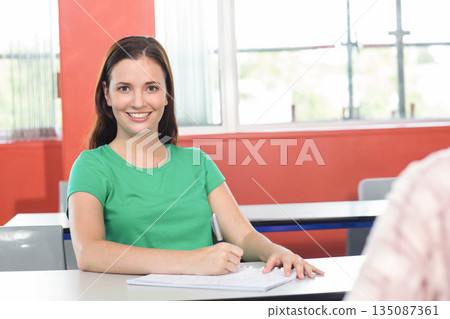 Female student sitting at classroom desk writing in notebook using ballpoint pen near window blinds 135087361