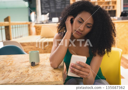 African American woman sitting at wooden cafe table scrolling smartphone and mug with tea bag near African American woman sitting at wooden cafe table scrolling smartphone and mug with tea bag near 135087362