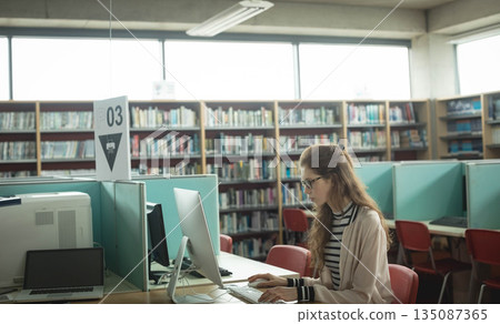 Teenage girl studying at library carrel using desktop computer and closed laptop, copy space 135087365