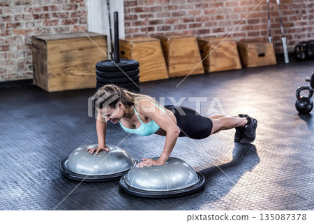 Woman wearing mint sports bra and leggings performing elevated push-ups on BOSU trainers at gym Woman wearing mint sports bra and leggings performing elevated push-ups on BOSU trainers at gym 135087378