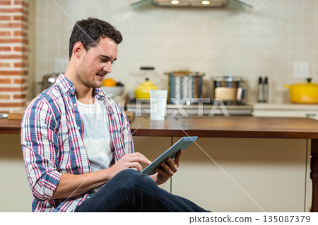 Man in his twenties sitting on floor using tablet in kitchen with coffee cup and pots Man in his twenties sitting on floor using tablet in kitchen with coffee cup and pots 135087379