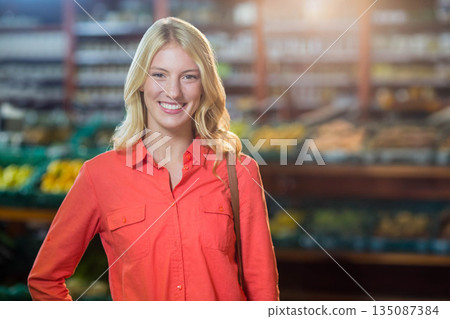 Female shopper standing in grocery store produce aisle examining produce bins and carrying tote bag 135087384