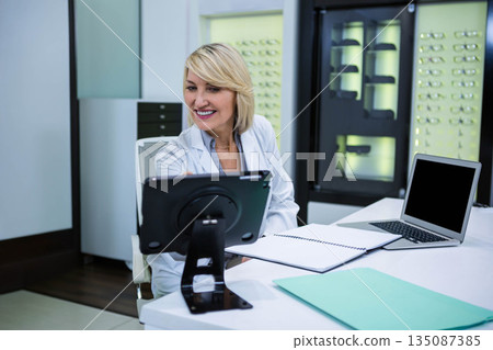 Senior woman wearing lab coat sitting at desk in optical clinic reviewing eyeglass frames on tablet Senior woman wearing lab coat sitting at desk in optical clinic reviewing eyeglass frames on tablet 135087385