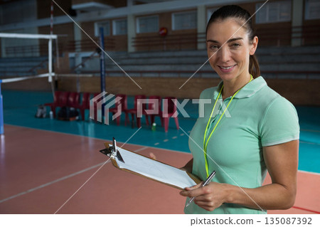 Female volleyball coach standing on court at gym holding clipboard and pen beside net, copy space 135087392