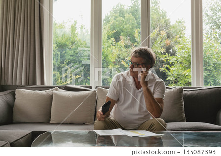 Senior man sitting on grey sofa holding remote and touching eyeglasses in living room, copy space Senior man sitting on grey sofa holding remote and touching eyeglasses in living room, copy space 135087393