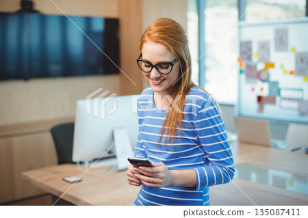 Red-haired woman standing by wooden office desk checking smartphone with monitor and whiteboard 135087411