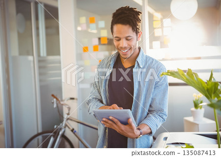 African American male tapping tablet near glass partition with sticky notes and bicycle in office 135087413