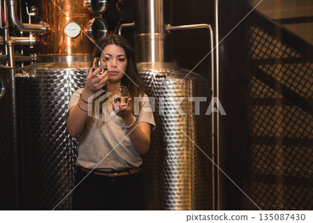 Adult woman technician holding and comparing two spirit samples in control room with steel tanks 135087430