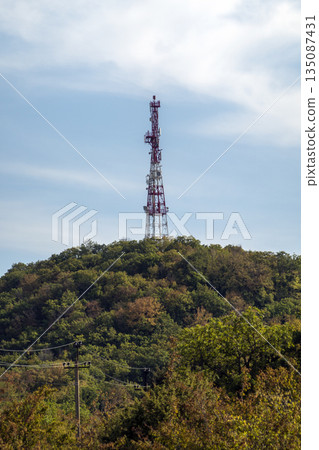 Red and white communication tower with multiple antennas standing on a mountain, representing telecommunications infrastructure, broadcasting technology and signal transmission Red and white communication tower with multiple antennas standing on a mountain, representing telecommunications infrastructure, broadcasting technology and signal transmission 135087431