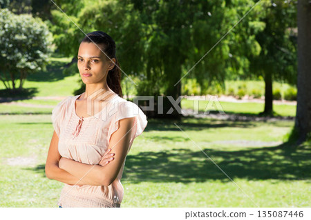Woman standing with crossed arms looking off camera in sunlit park with green grass, copy space 135087446