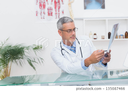 Male doctor examining x-ray film while sitting at glass desk in consultation room with stethoscope 135087466