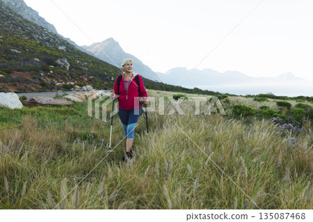 Woman hiking using trekking poles on grassy plain at mountain foothills wearing red jacket boots Woman hiking using trekking poles on grassy plain at mountain foothills wearing red jacket boots 135087468