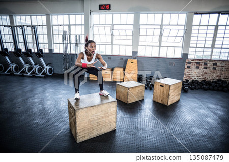 African American woman performing box jump in sportswear at CrossFit gym with clock, copy space African American woman performing box jump in sportswear at CrossFit gym with clock, copy space 135087479