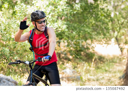 Mature male cyclist red sleeveless jersey riding black bike on forest trail carrying hydration pack 135087492