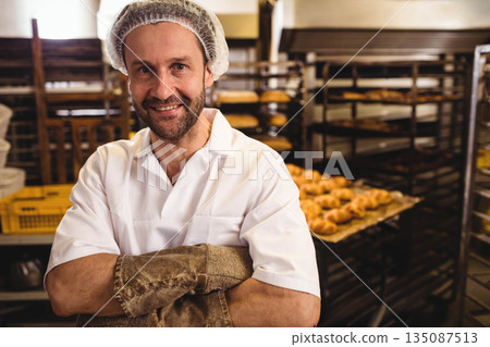 Male baker standing in bakery area wearing hairnet, leather oven gloves before metal racks of buns 135087513