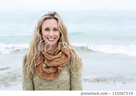 Woman standing on beach smiling at camera wearing olive green sweater with textured brown scarf Woman standing on beach smiling at camera wearing olive green sweater with textured brown scarf 135087519