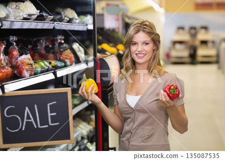Female shopper holding yellow and red bell peppers in supermarket produce aisle with sale sign 135087535