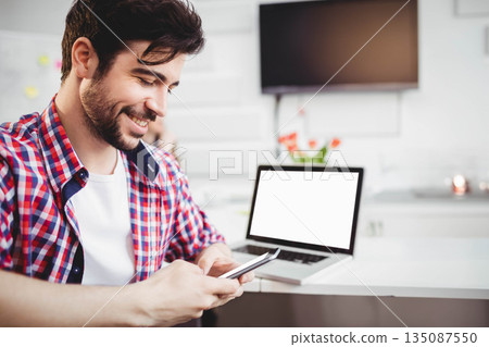 Man smiling while checking smartphone at home workspace with laptop and tulip vase, copy space 135087550