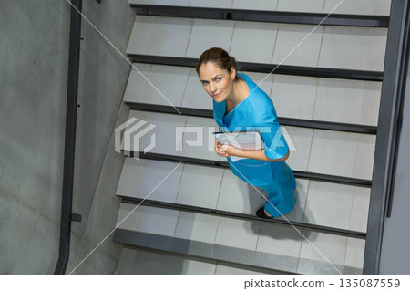 Mid-adult female medical worker in scrubs standing on tiled stairwell in hospital holding clipboard 135087559
