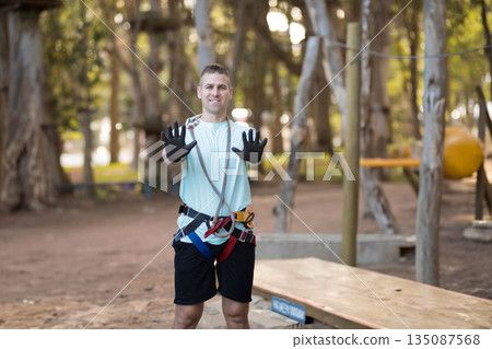 Middle-aged man wearing harness and gloves standing on low platform among ropes in park 135087568