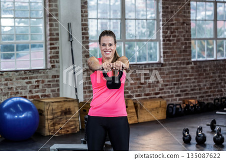 Mid adult woman wearing pink tank top lifting kettlebell in industrial gym with stability ball 135087622